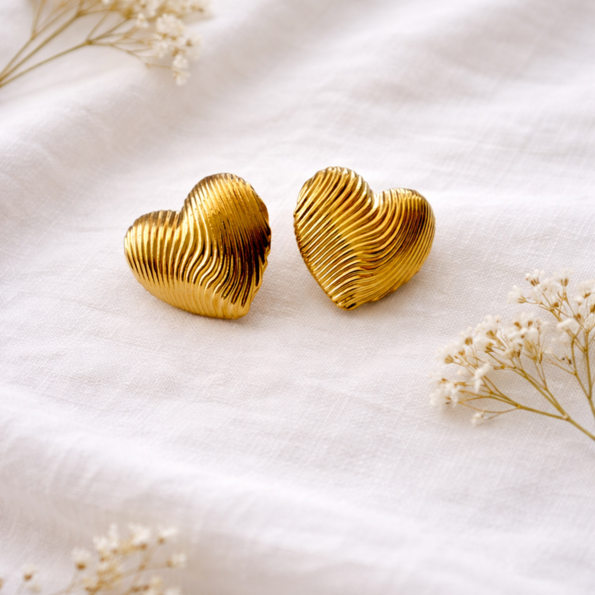 Paire de boucles d'oreilles puces en forme de cœur bombé doré en or fin 24k, avec motif de stries ondulées, posées sur un tissu blanc entouré de fleurs séchées