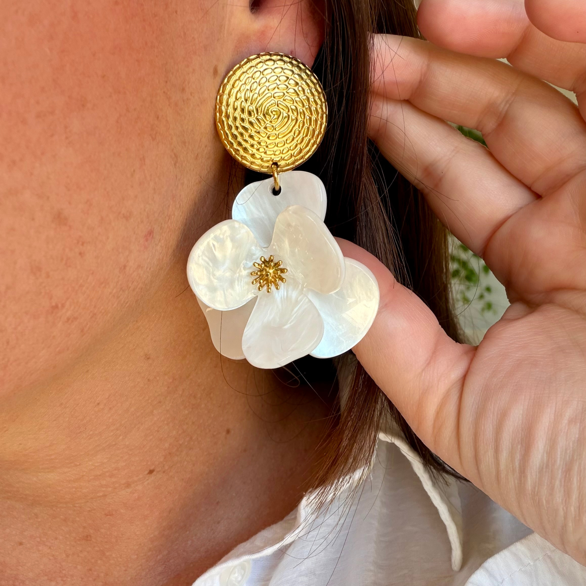 Boucle d'oreille pendante portée de profil — puce ronde en acier doré texturé spirale, pendentif fleur en résine nacrée blanche aux pétales arrondis avec pistil doré central, sur chemise blanche