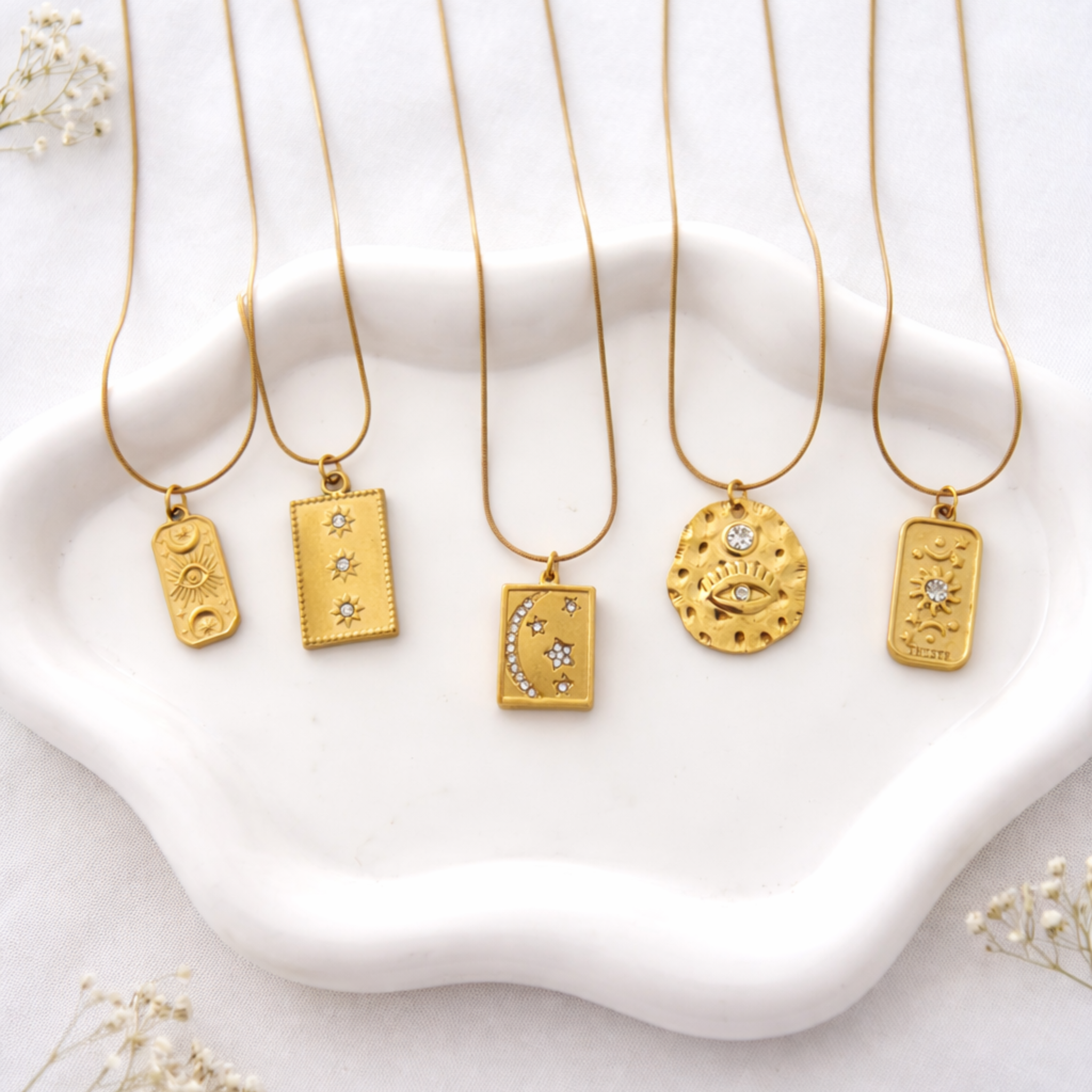 Collection de 5 colliers talisman dorés en acier inoxydable avec pendentifs œil, lune, soleil et étoiles posés sur plateau blanc